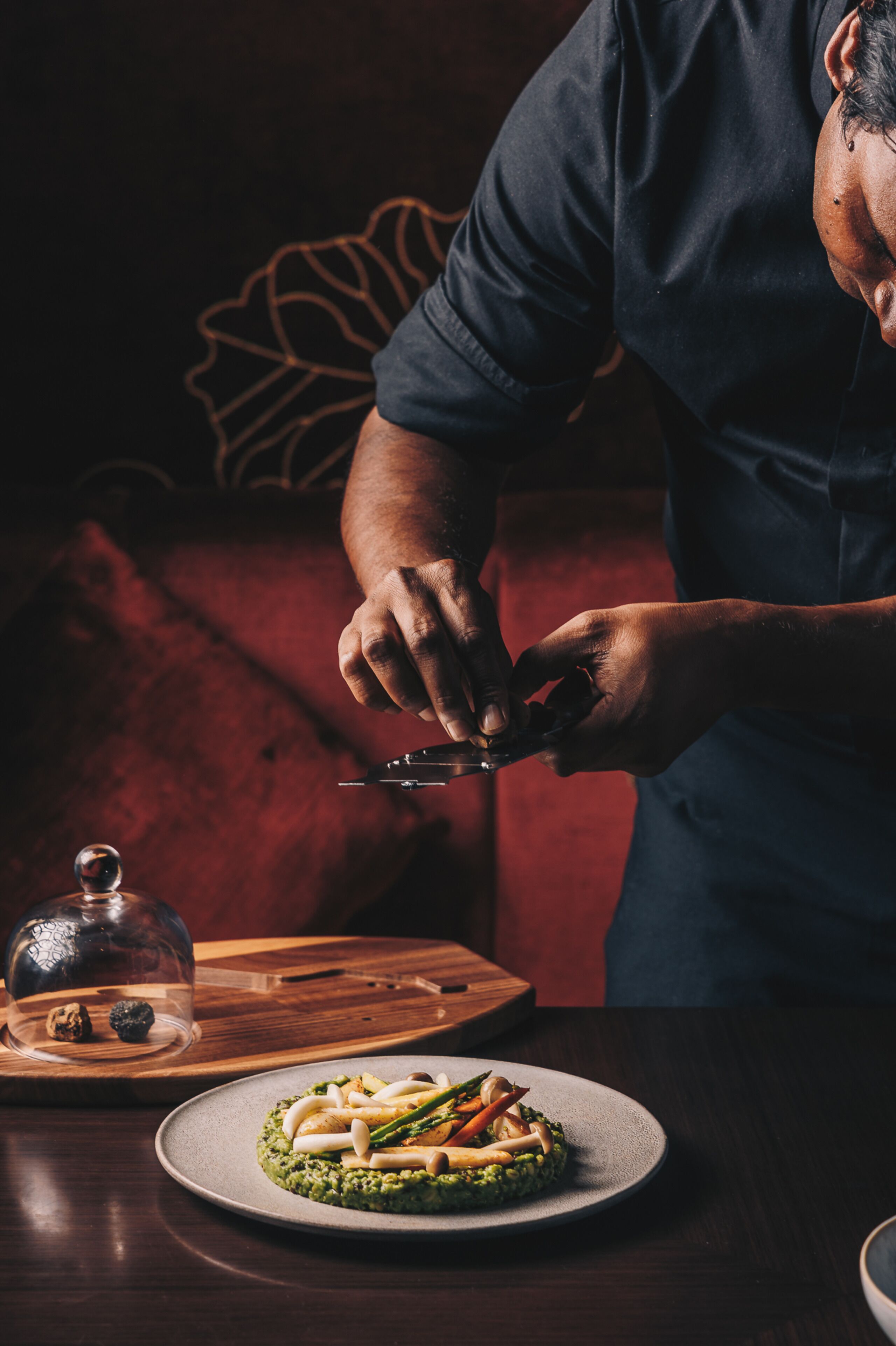 Une personne découpe un plat sur une table en bois, et un cloche de verre contenant des noix est posée à proximité.