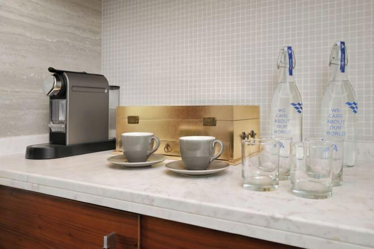 A kitchen counter with a coffee maker, two cups, a glass carafe, and two bottles with labels.
