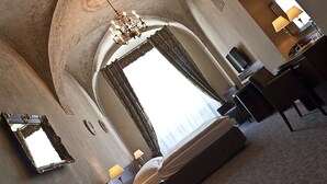 A hotel room with a large bed, a chandelier, a desk with a chair, and a large window with curtains.