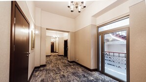 A hotel hallway with patterned carpet, a chandelier, and a large glass door with a view of a building.