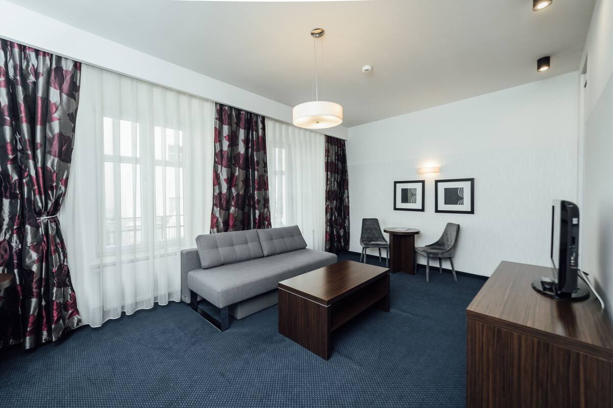 Una habitación de hotel moderna con un sofá gris, una mesa de centro de madera, un escritorio con una computadora, dos cuadros en la pared y un ventanal con cortinas traslúcidas.