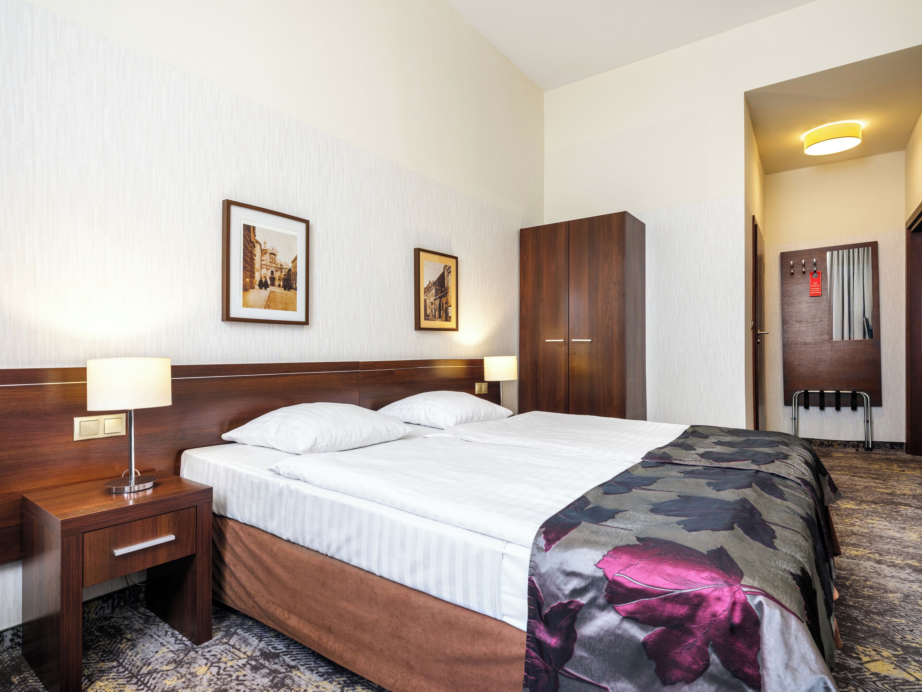 Habitació d'hotel amb un llit, un panell de capçalera de fusta, dues taules de nit amb làmpades, un armari i dues imatges emmarcades a la paret.
