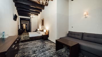 A hotel room with a bed, a sofa, a wooden coffee table, a patterned rug, a chandelier, and a wall-mounted lamp.