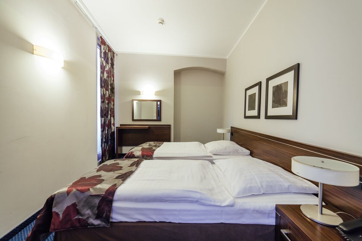 A hotel room with two beds, a desk, a chair, a lamp, a mirror, and two framed pictures on the wall.