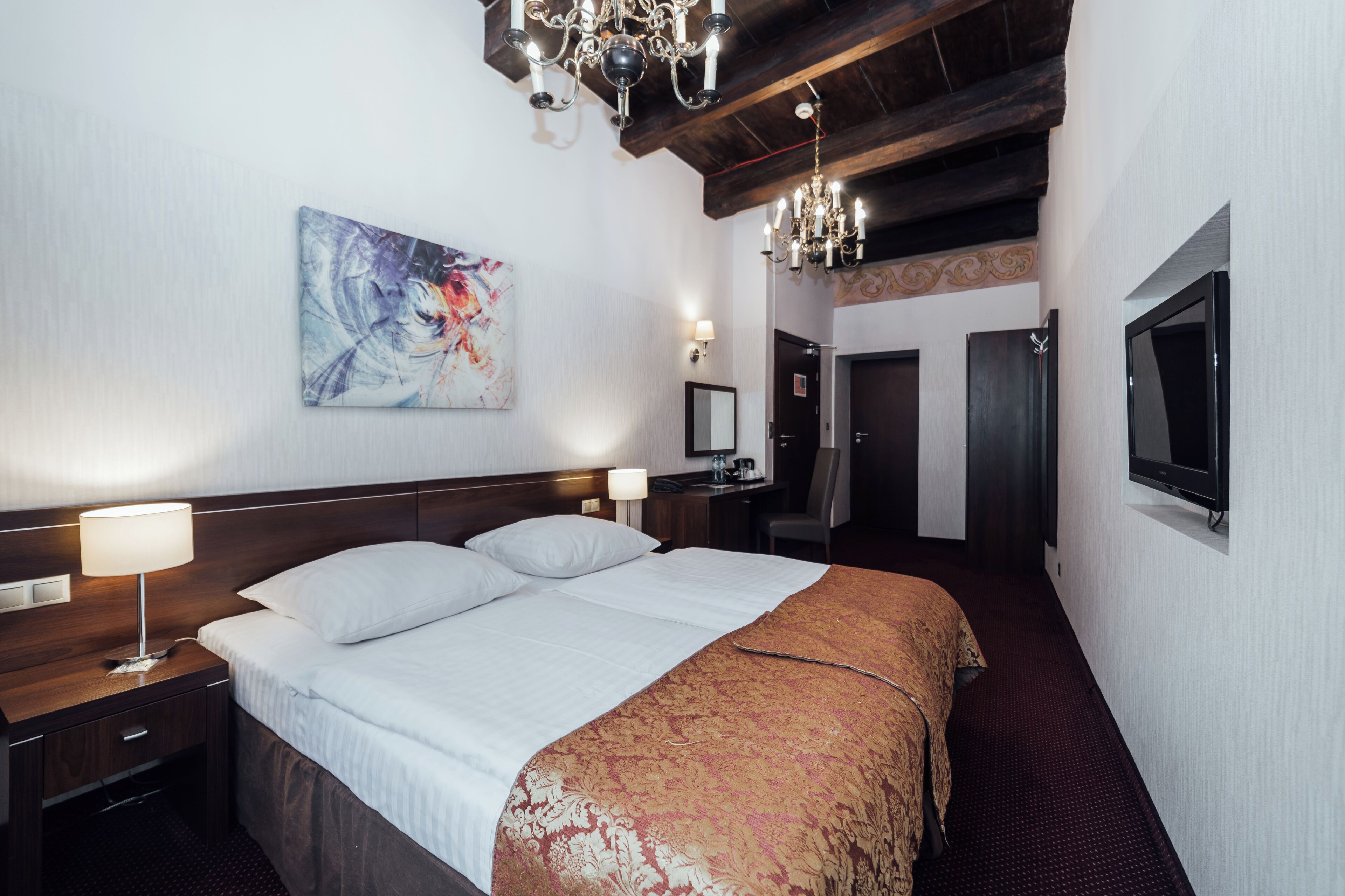 A hotel room with a large bed, a desk with a lamp, a television, and a painting on the wall.