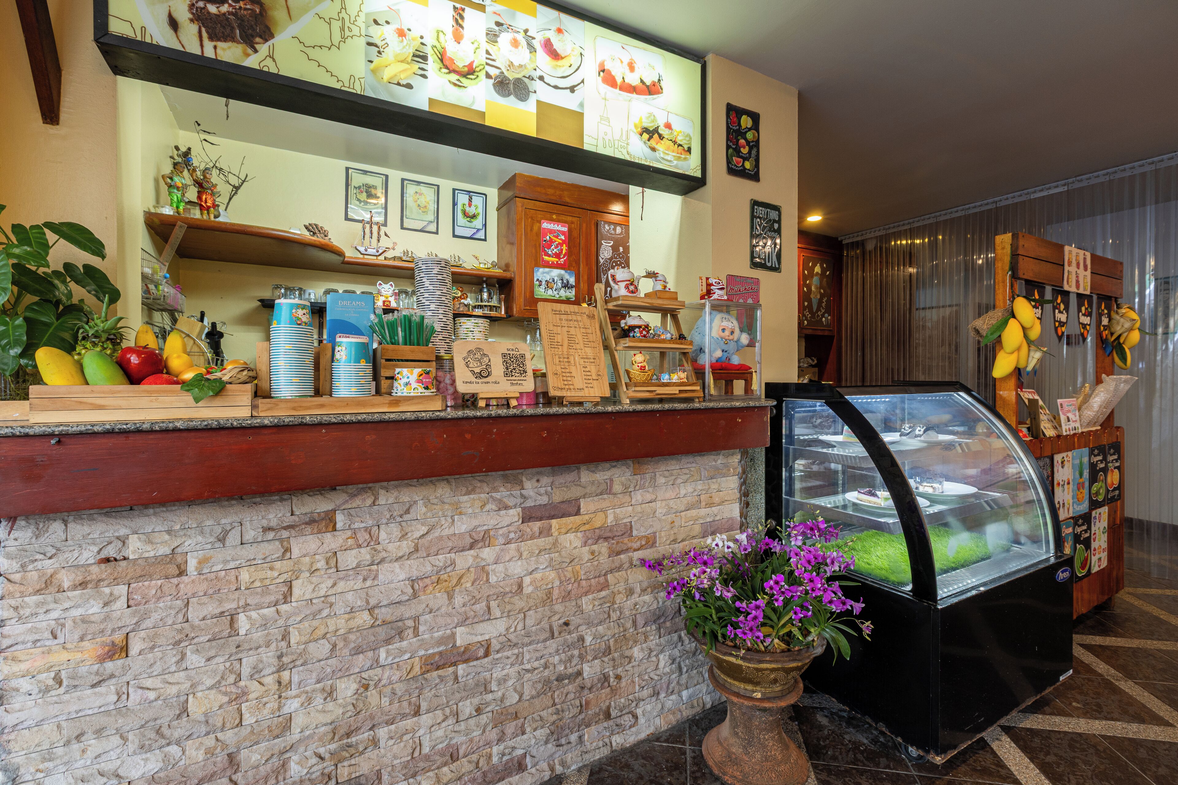 Un hall d’accueil d’hôtel avec un mur en pierre, un présentoir de fruits, une vitrine avec des fleurs et un comptoir en bois où sont exposés divers objets.