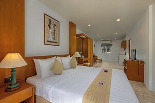 A hotel room with a large bed, wooden headboard, a nightstand with a lamp, a desk with a chair, a mirror, and a television.