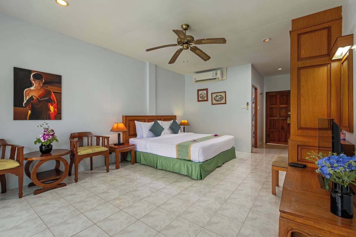 A hotel room with a bed, two chairs, a desk, a ceiling fan, and a wall-mounted TV.