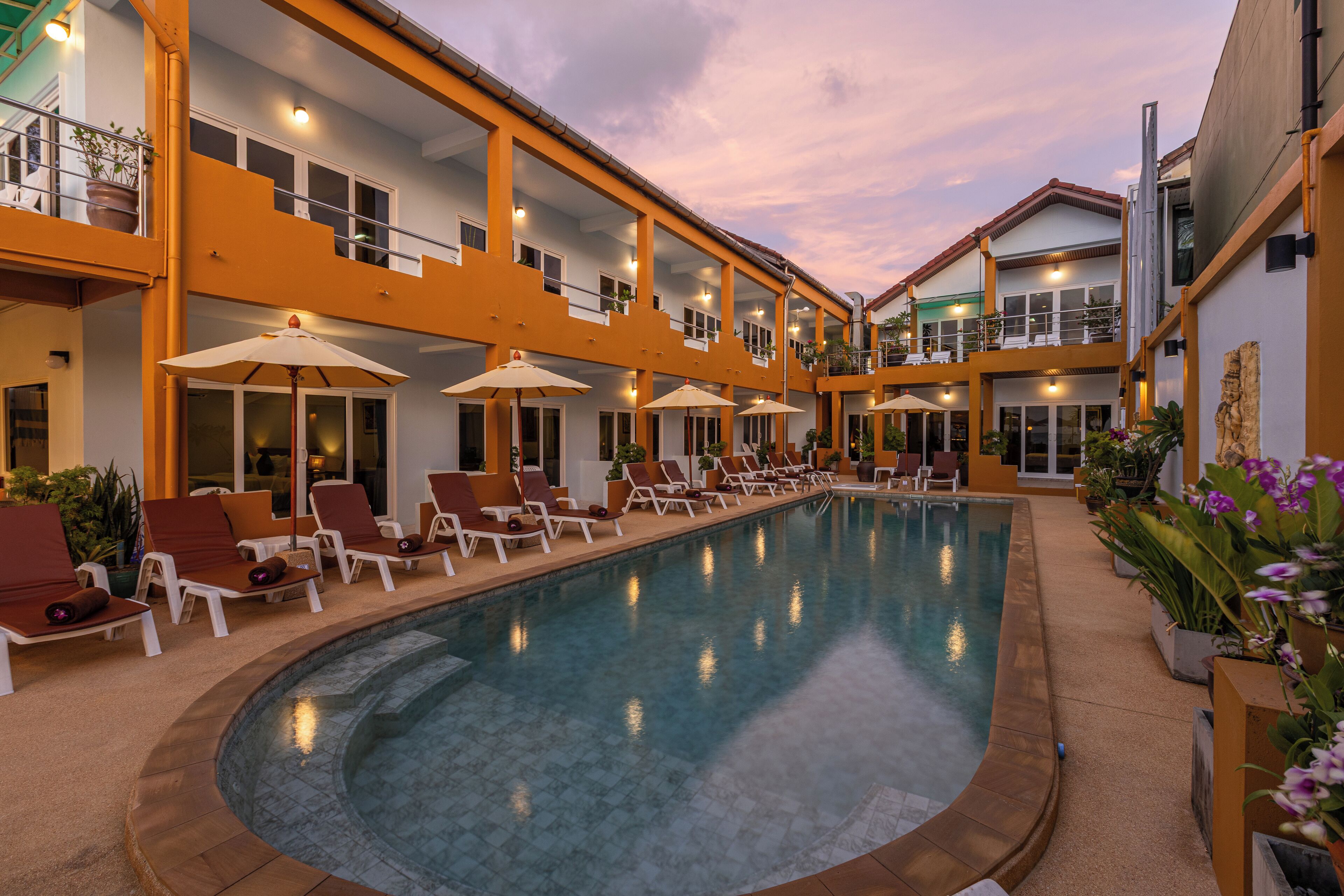 A pool area with lounge chairs and umbrellas, a multi-story building with balconies, and a clear sky.
