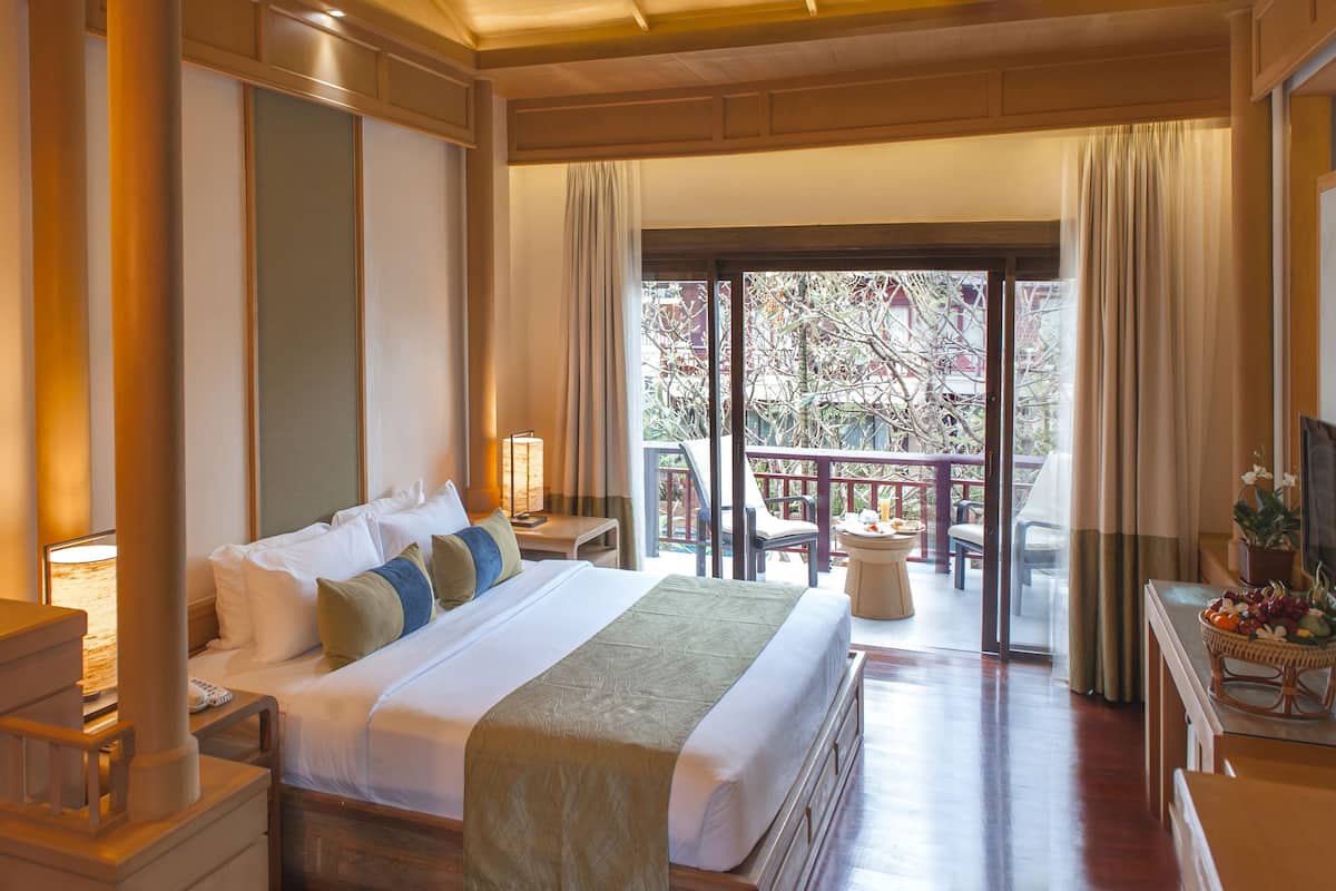 Habitación de hotel con una cama grande, una mesita de noche, una lámpara, una pequeña mesa con una cesta de fruta y vistas al exterior.