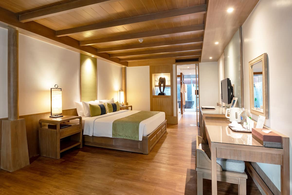 Habitación de hotel con techo de madera, cama con manta verde, mesita de noche con lámpara, escritorio con espejo y televisor.