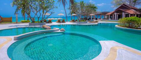 A swimming pool with a curved design, surrounded by lounge chairs and umbrellas, with a view of the ocean and a building in the background.