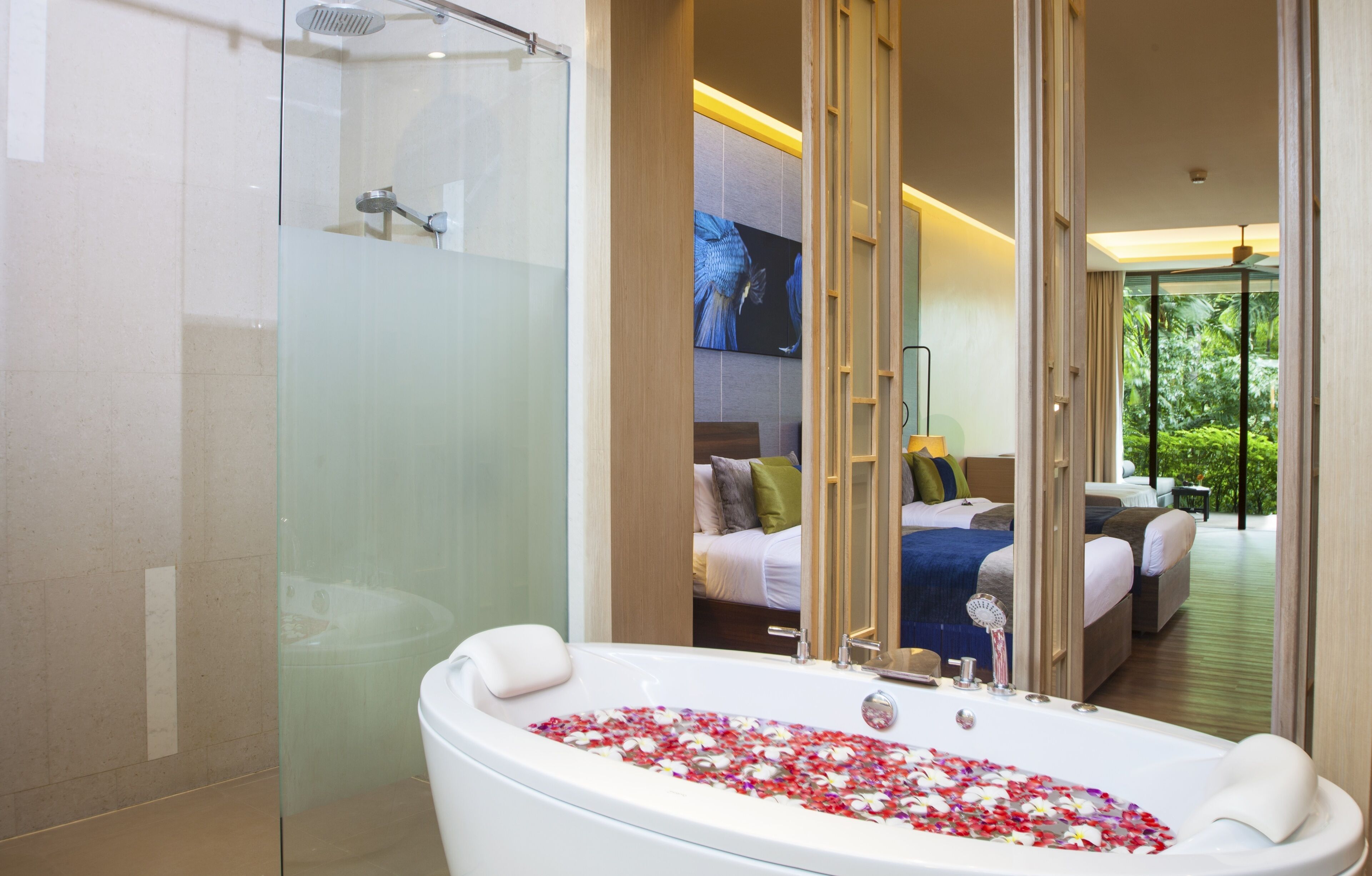A modern bathroom with a glass-enclosed shower, a bathtub filled with rose petals, and a bedroom visible through a mirrored wall.