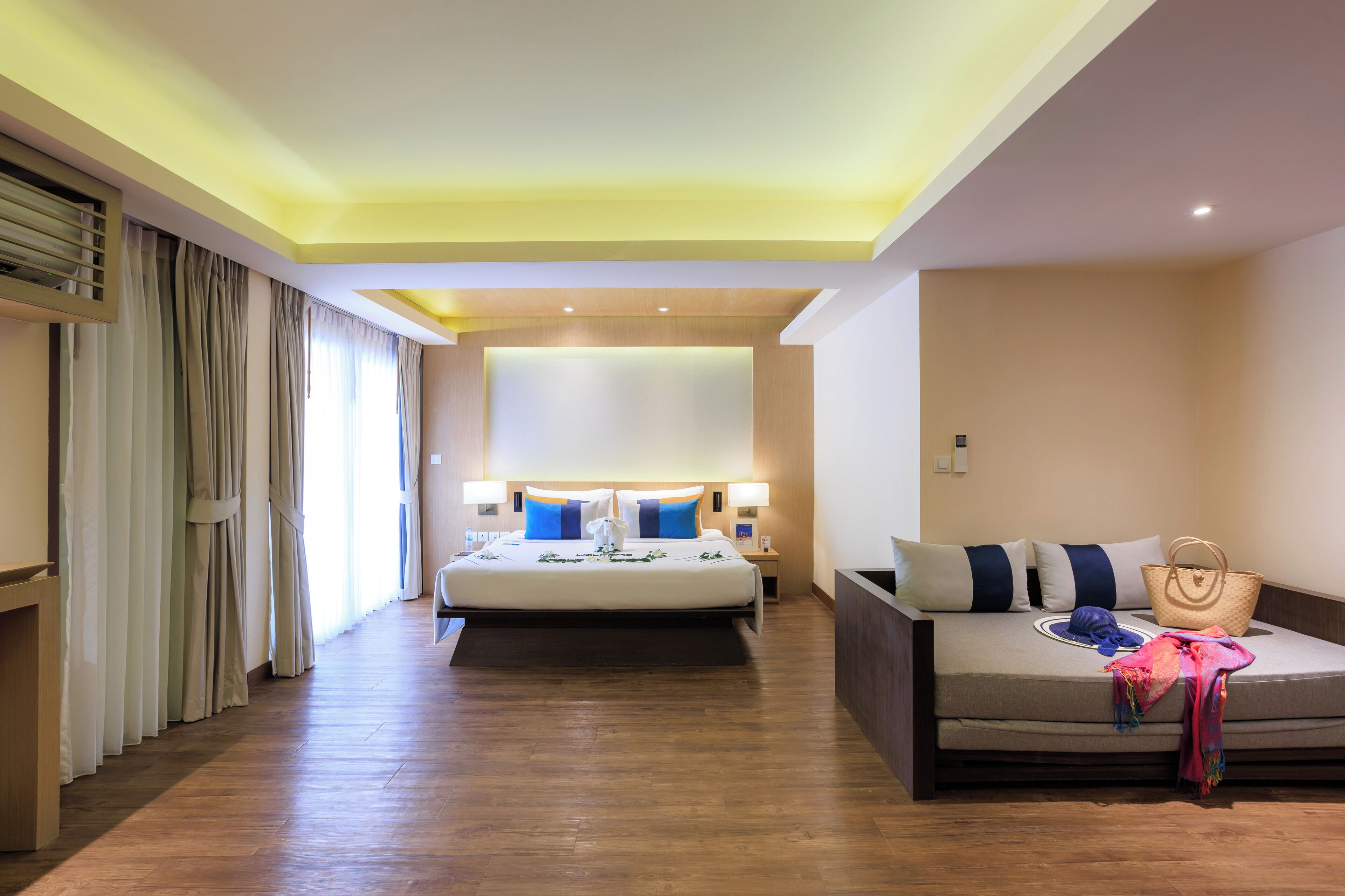 A hotel room with a bed, a sofa, a bedside table, a lamp, and a large window with curtains.