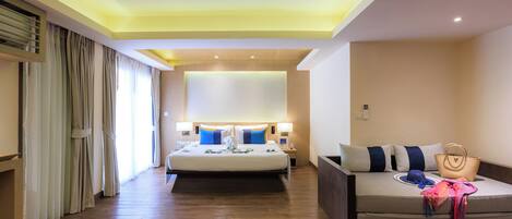 Habitación de hotel con cama, sofá, mesita de noche, lámpara y un gran ventanal con cortinas.