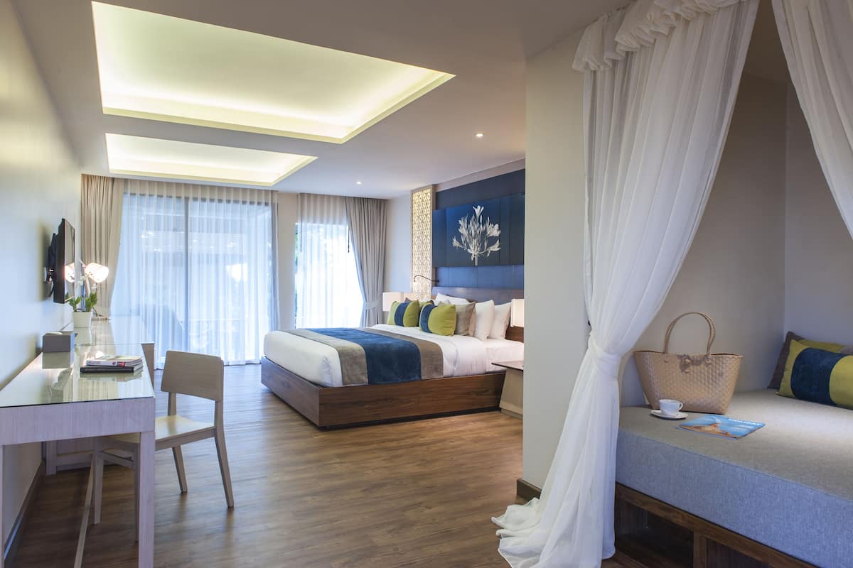 Una habitación de hotel moderna con una cama grande, un escritorio con silla, una mesita de noche y un amplio ventanal con cortinas.