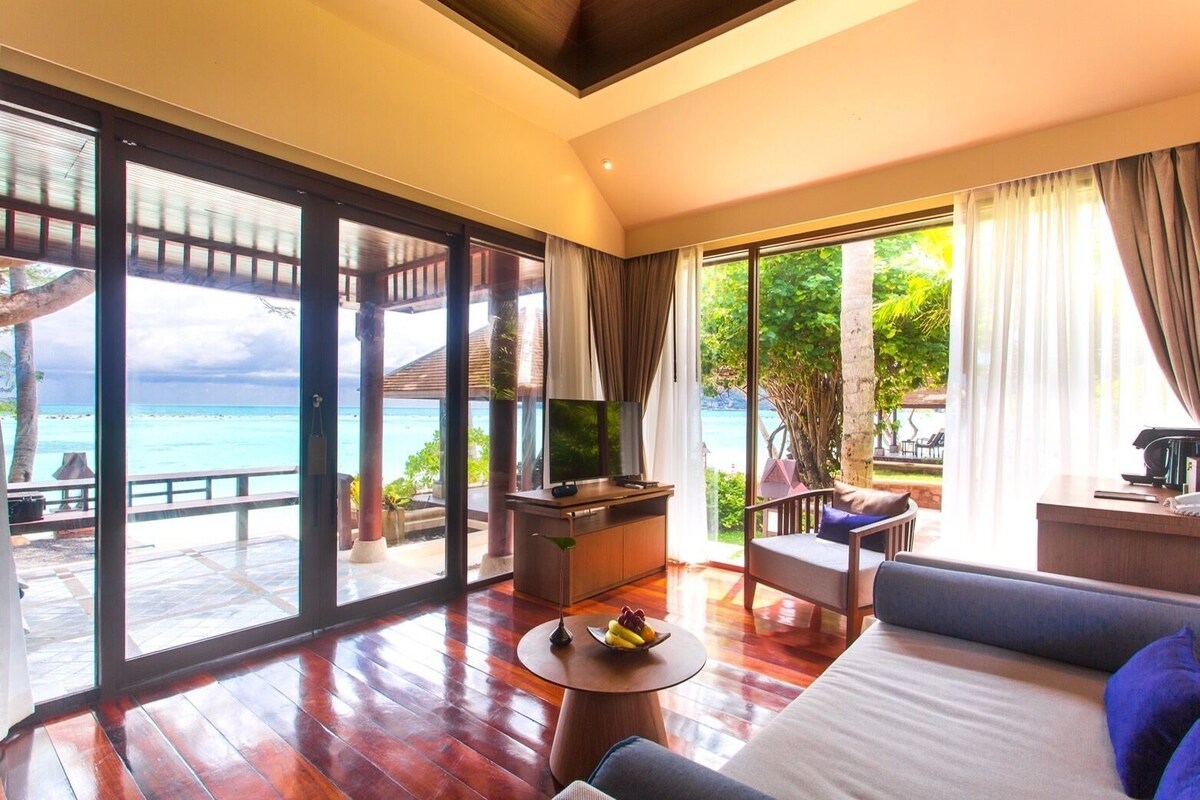 A spacious room with a large sliding glass door leading to a balcony with a view of the ocean. There is a sofa, a small table with fruit, and a television on a wooden cabinet.