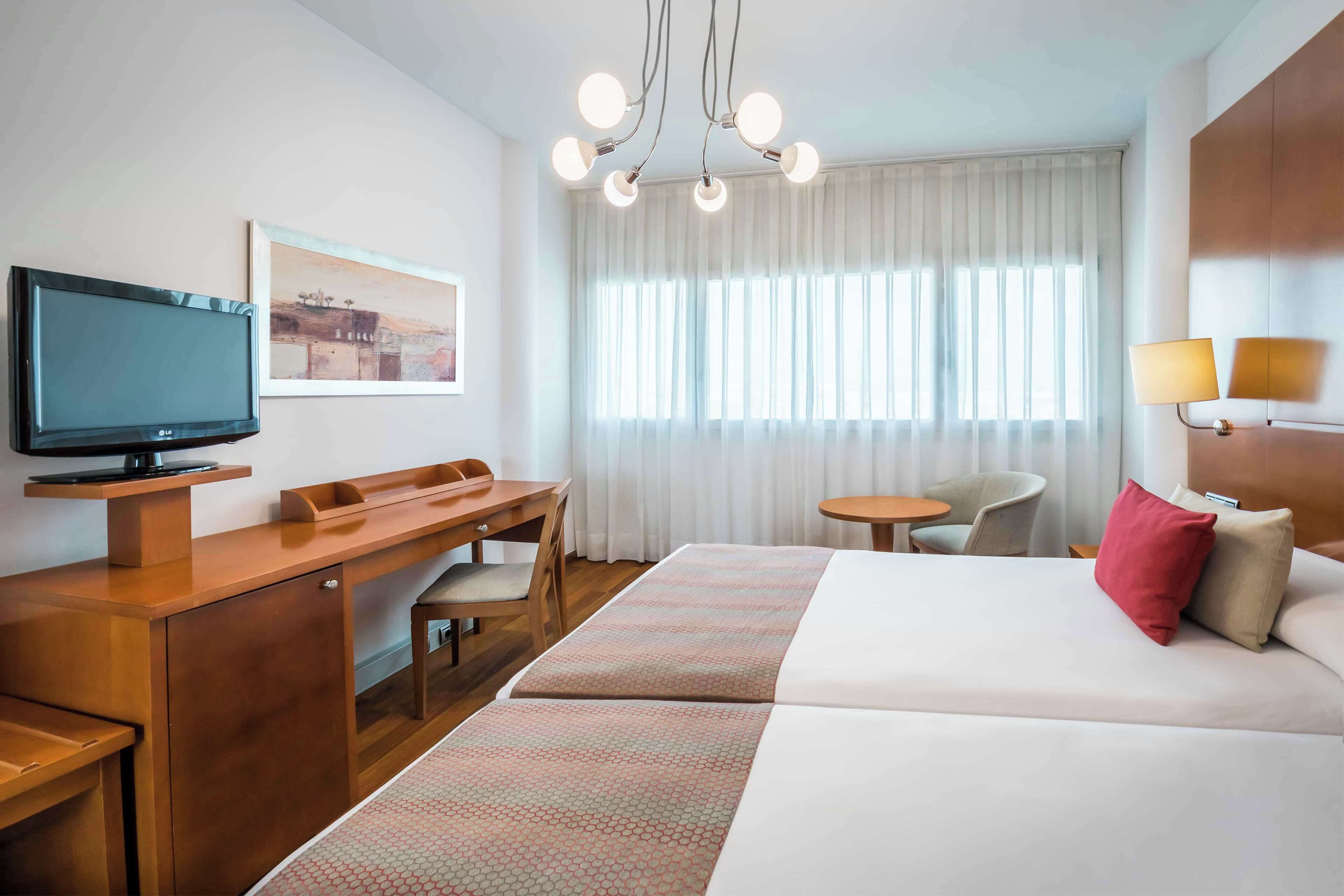 A hotel room with a bed, a desk, a chair, a television, a lamp, and a window with curtains.
