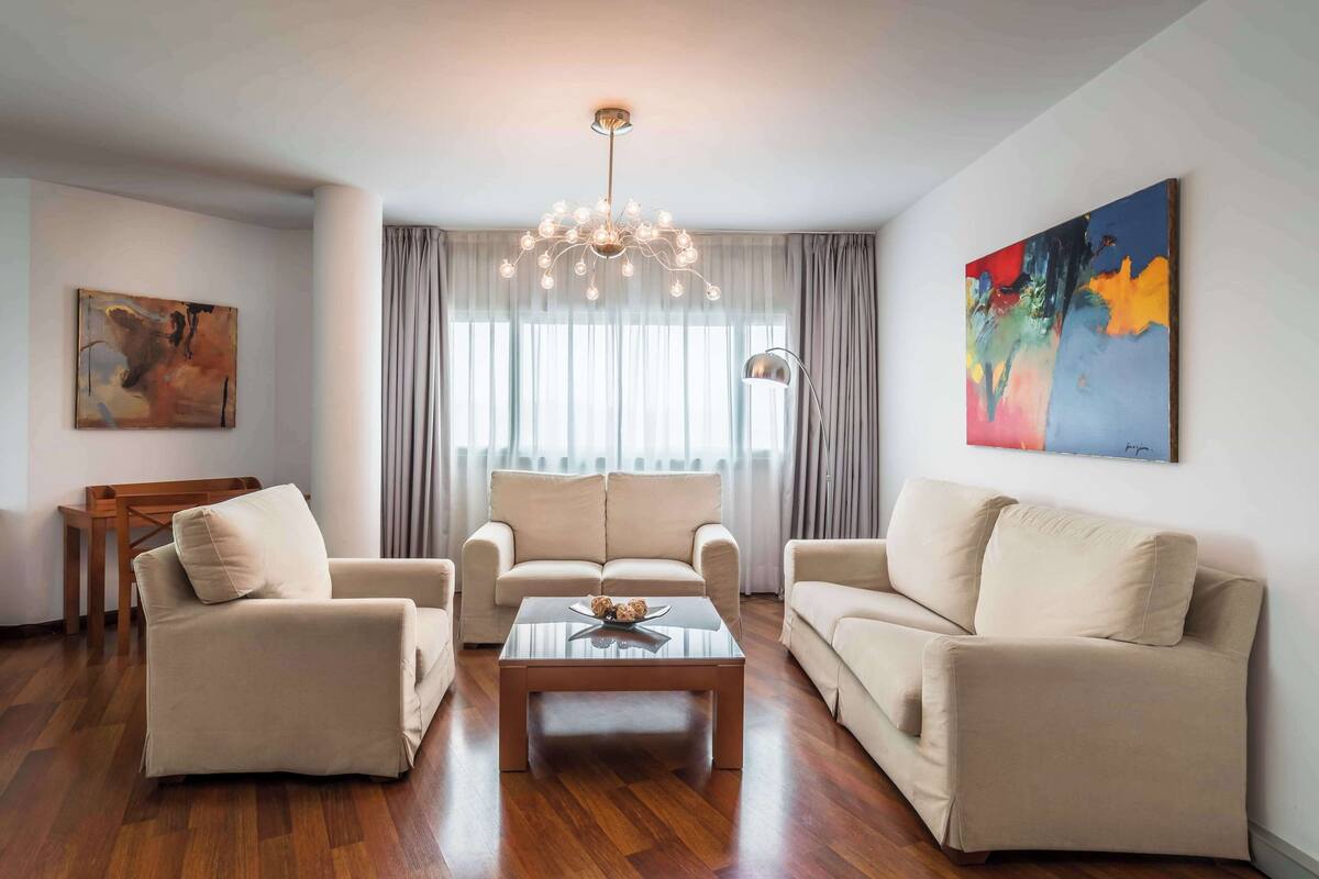 A living room with a beige sofa set, a glass coffee table, a chandelier, and a painting on the wall.