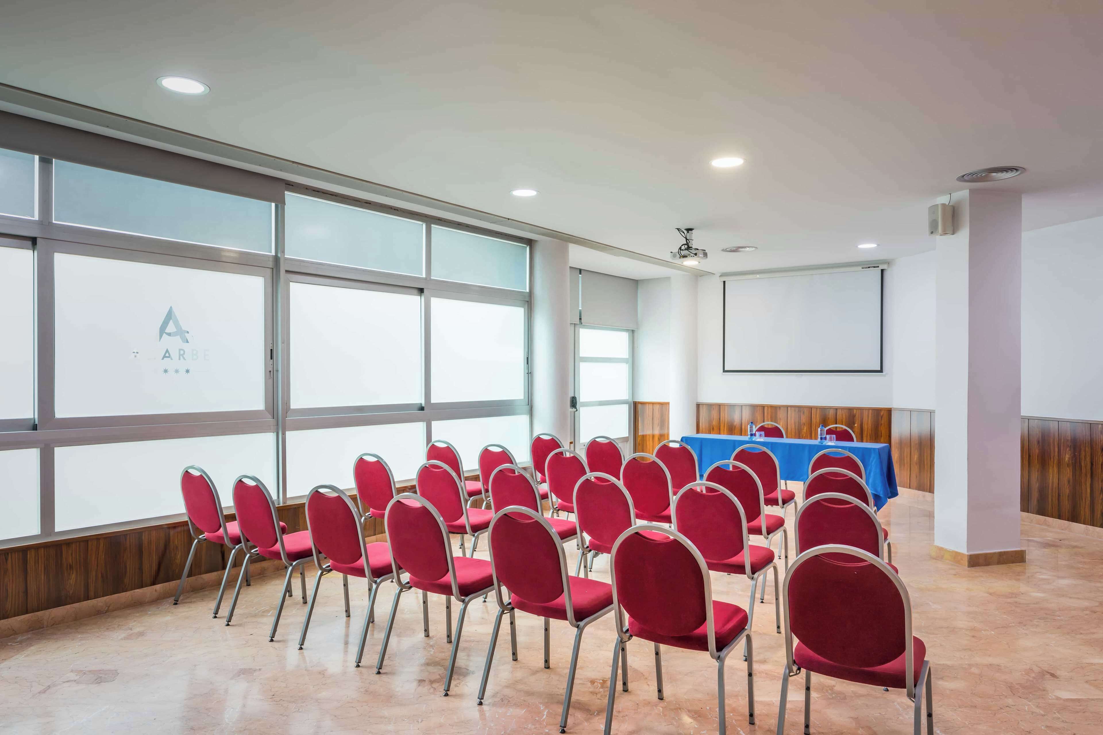 Un salón de conferencias con hileras de sillas rojas, una mesa azul y una pantalla de proyector.