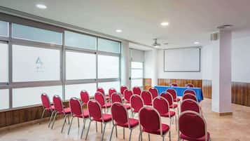 Un salón de conferencias con hileras de sillas rojas, una mesa azul y una pantalla de proyector.