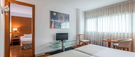 Habitación de hotel con cama, televisor sobre una mesa de vidrio, una silla, una mesita y una ventana con cortinas.