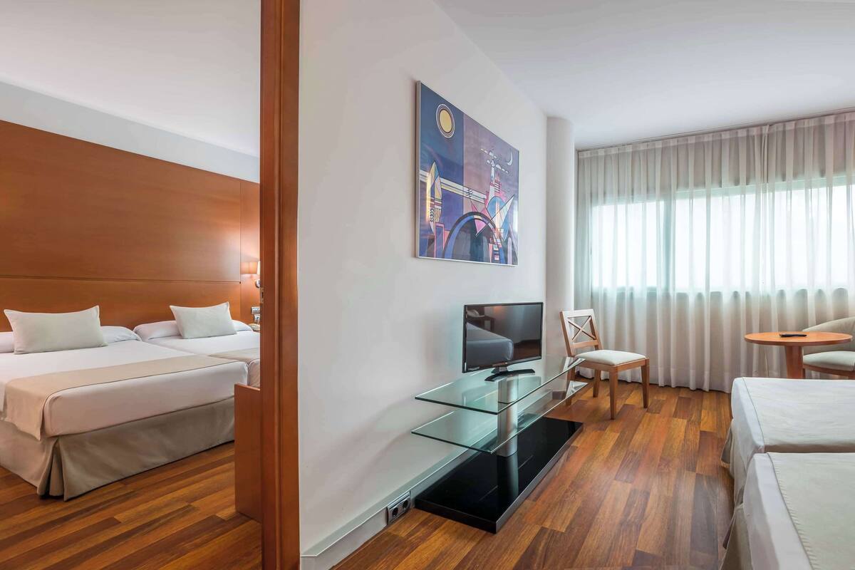 A hotel room with a bed, a desk with a TV, a chair, and a window with curtains.