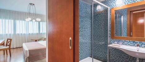 Habitación de hotel con cama, una silla, escritorio y baño con lavamanos y ducha.