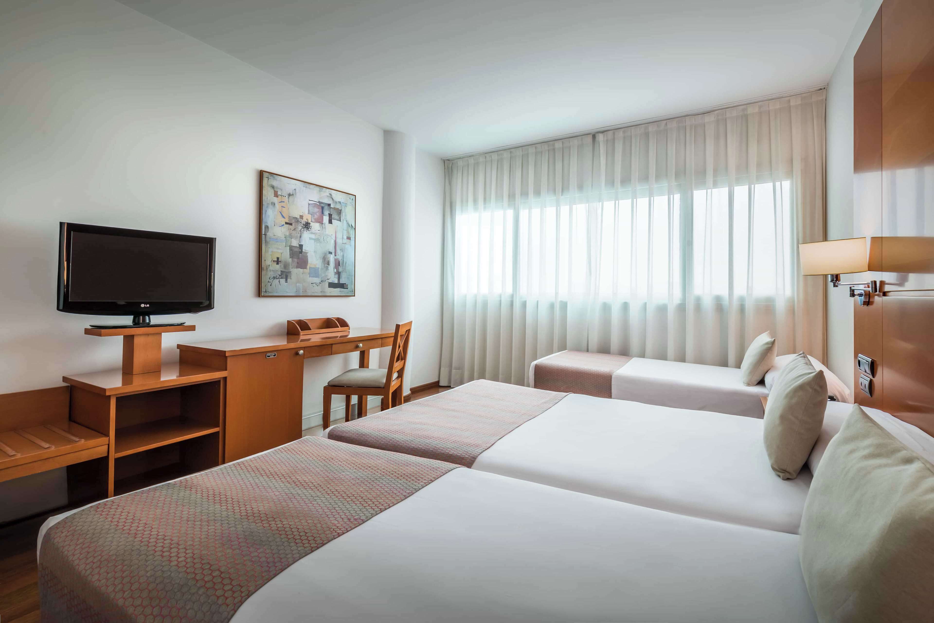 Habitación de hotel con dos camas, un escritorio, televisor y ventana con cortinas.
