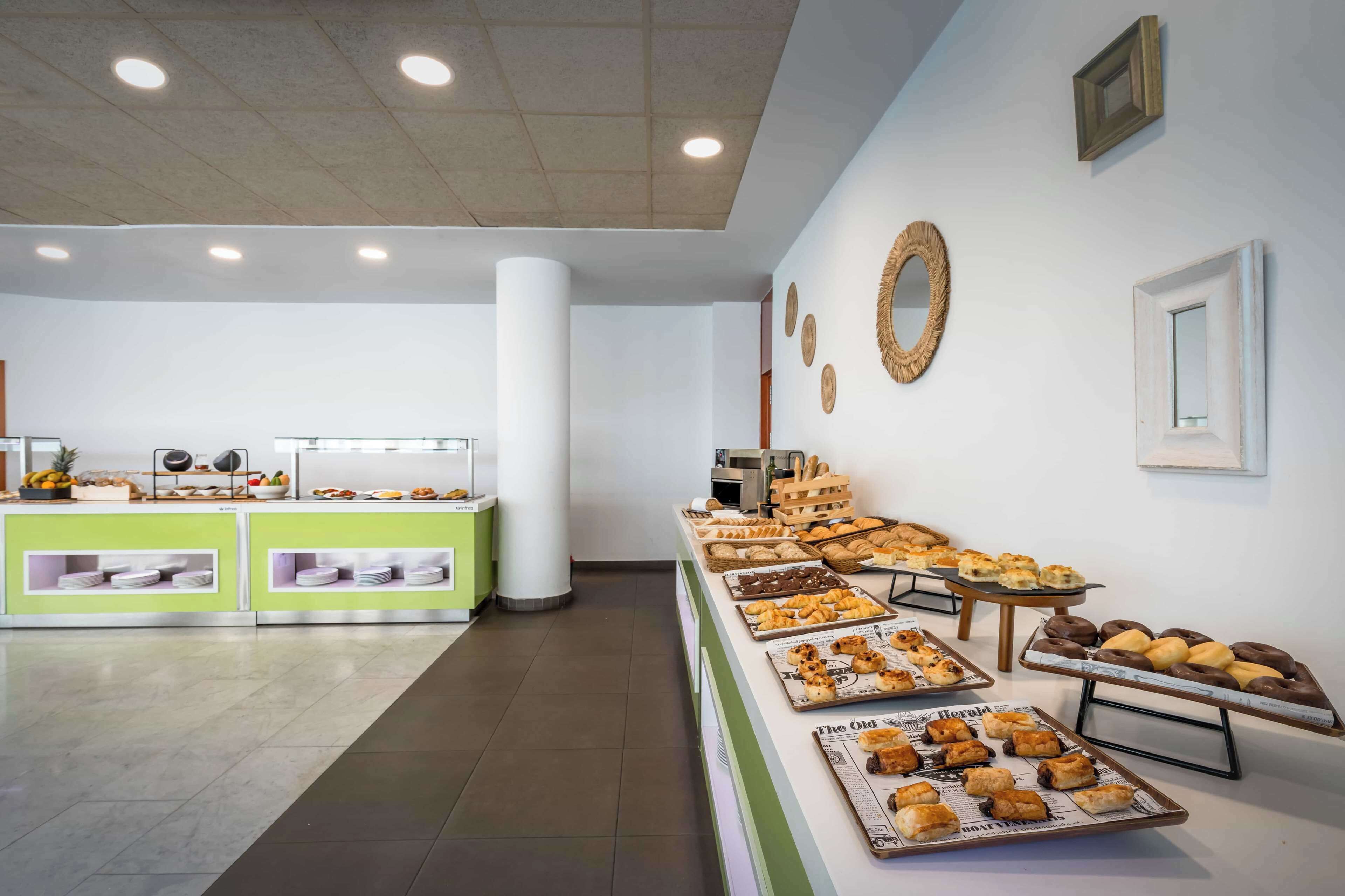 Zona de bufé con diversos alimentos expuestos en bandejas, una barra con sección de frutas y verduras, y una pared decorada.