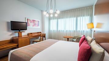 Ett hotellrum med en säng, ett skrivbord, en tv, en stol, en lampa och ett fönster med gardiner.