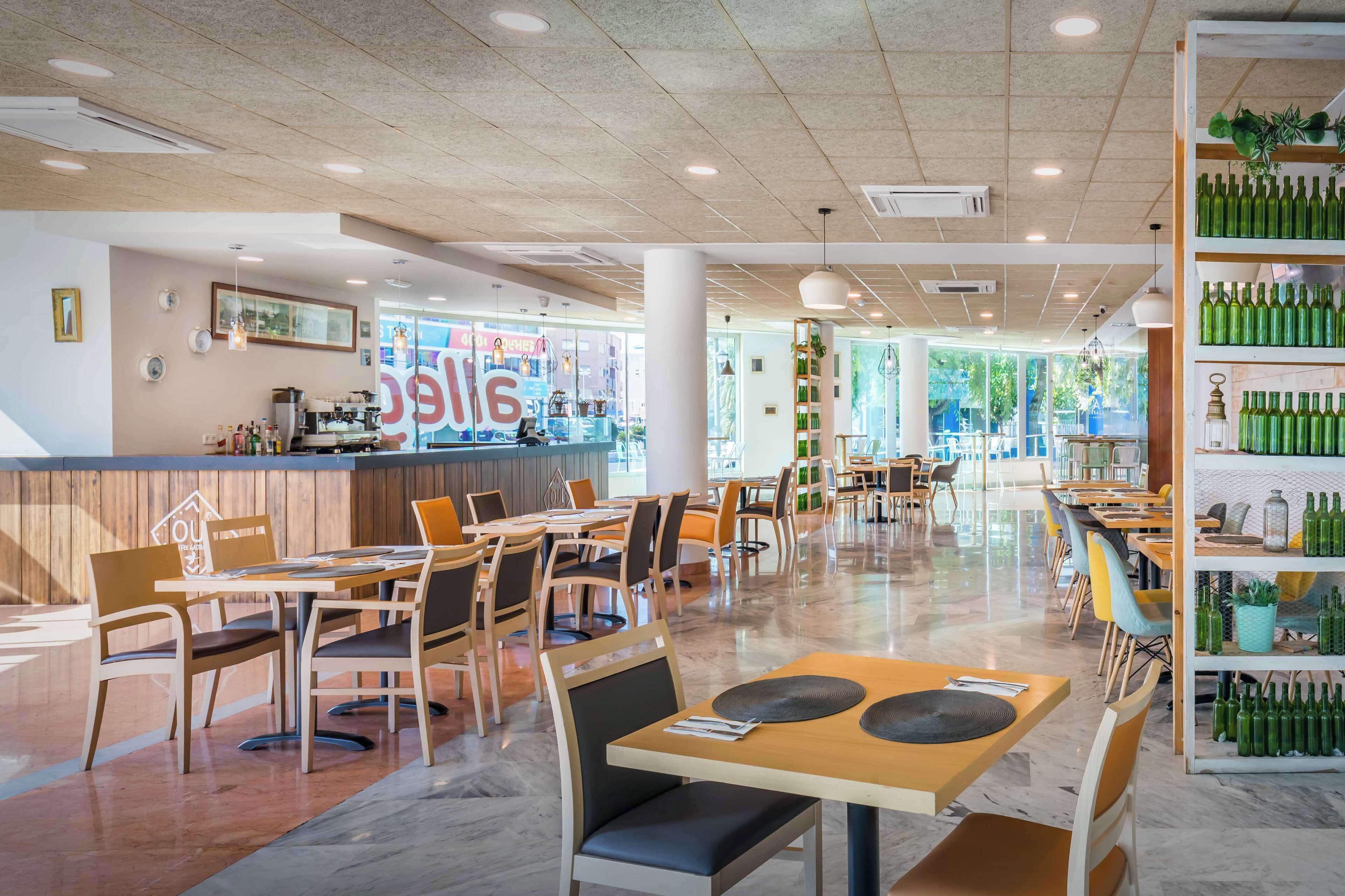 A spacious restaurant with wooden tables and chairs, a bar area, and a shelf with green bottles.