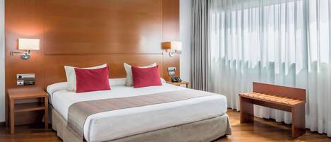 A hotel room with a large bed, two bedside tables with lamps, a bench, and a large window with curtains.