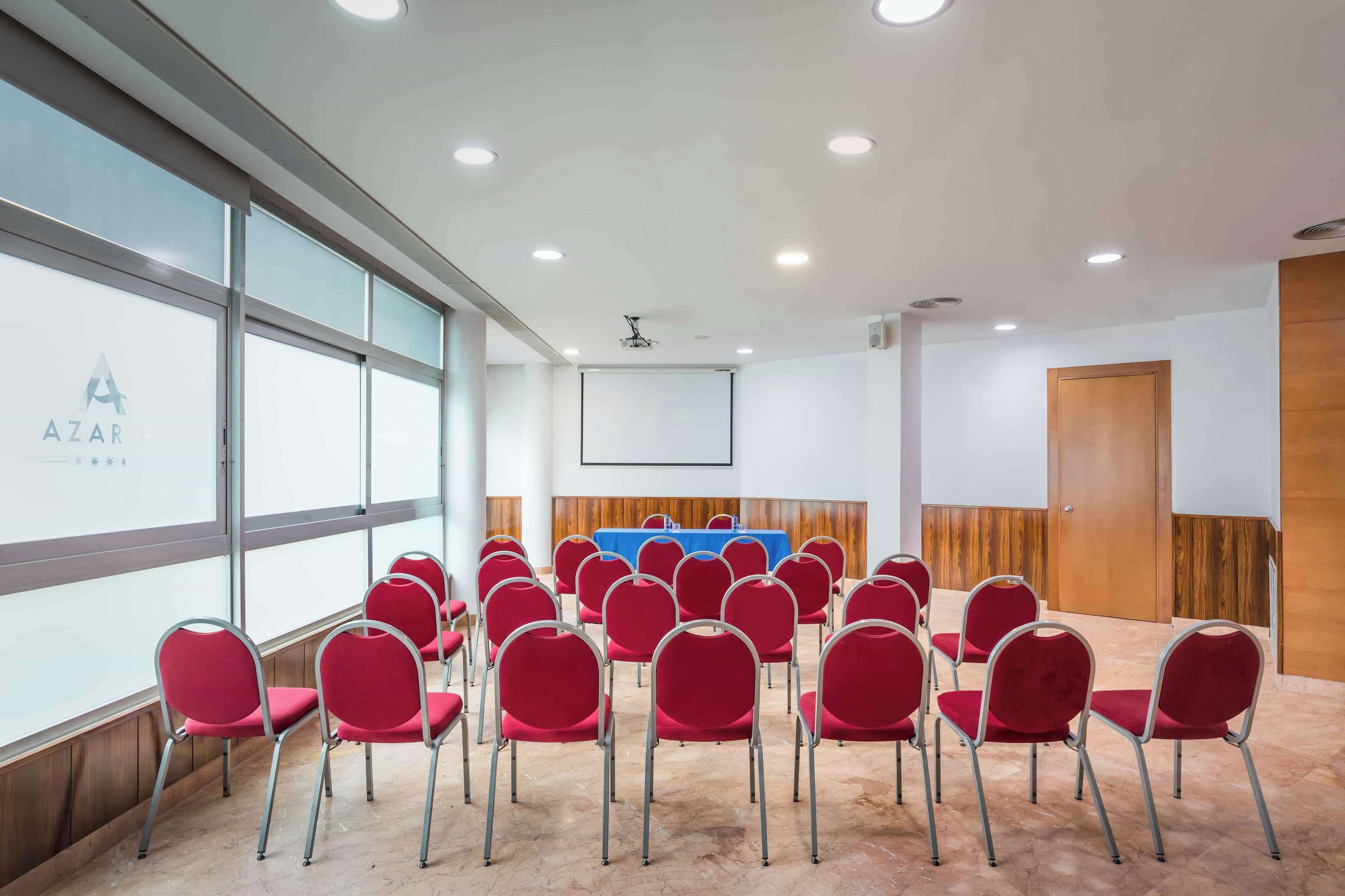 Een vergaderzaal met rode stoelen in rijen, een blauwe tafel en een projectiescherm.