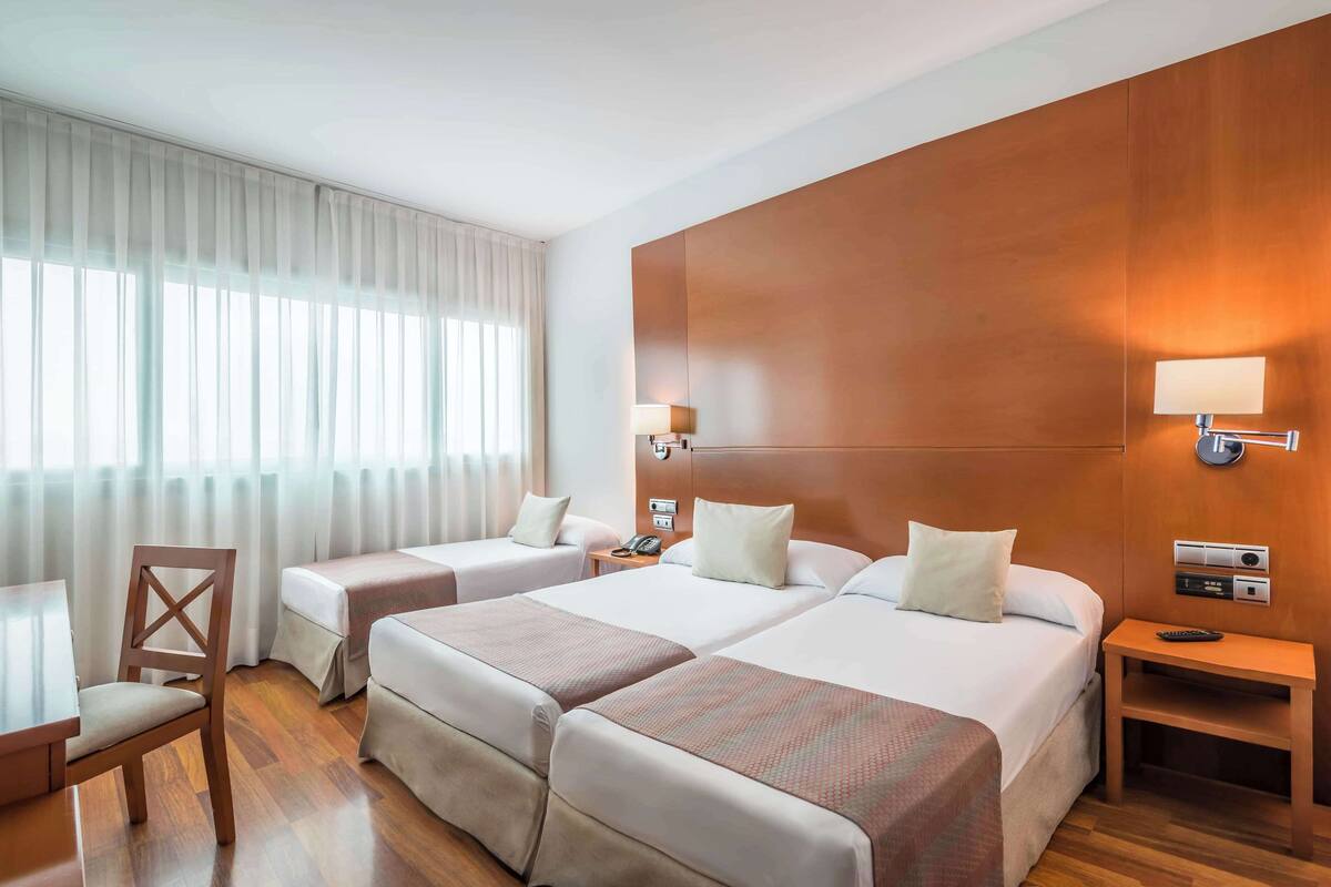 A hotel room with two beds, a desk, a chair, and a nightstand with a lamp.