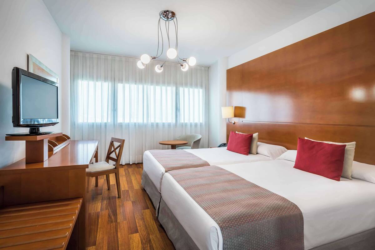 A hotel room with a wooden desk, a television, a bed with red pillows, a small table, and a chair.