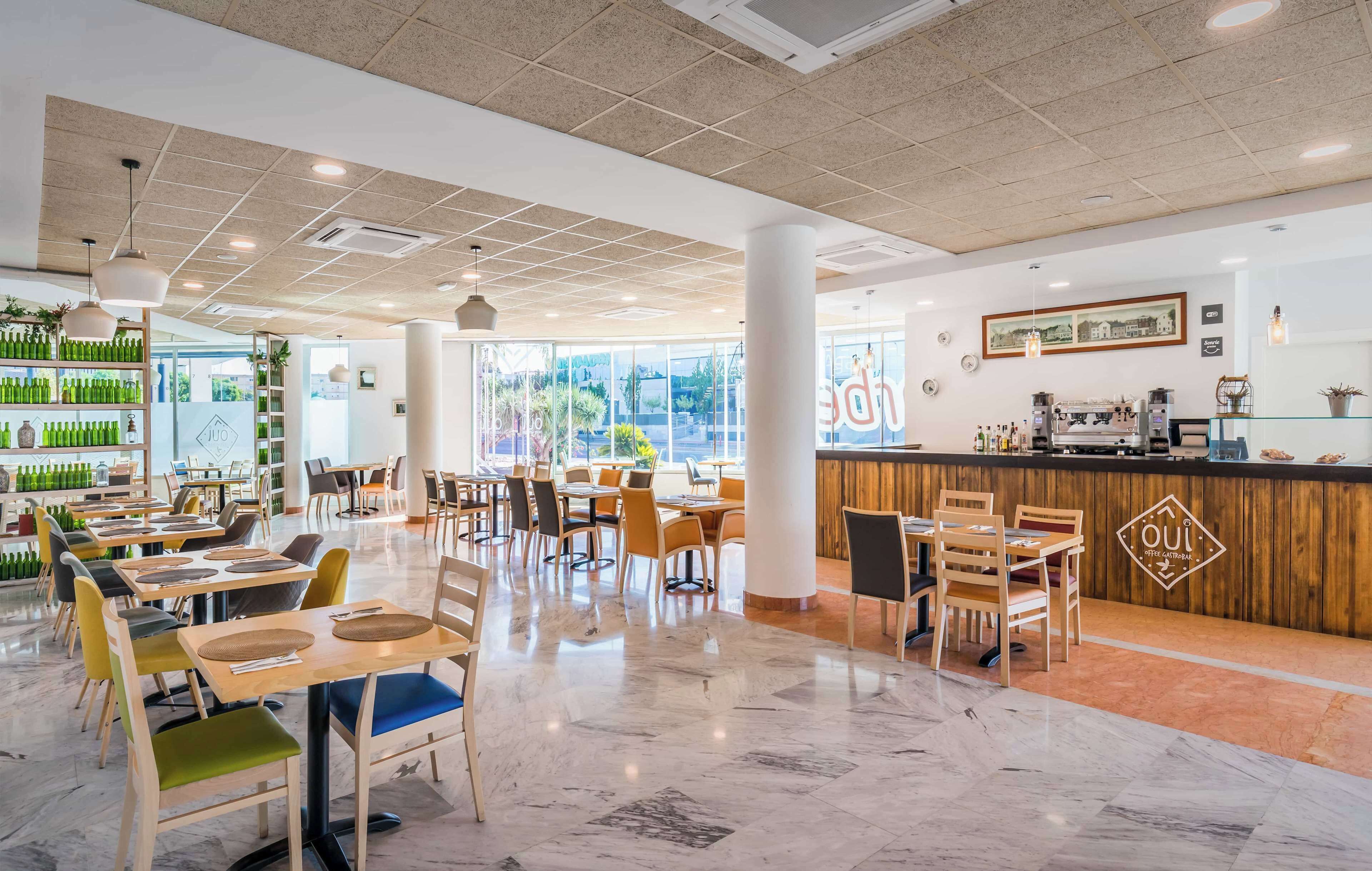 Een modern restaurant met een strak, open interieur, een centrale bar, verschillende zithoeken en een uitzicht op het buitenleven door grote ramen.