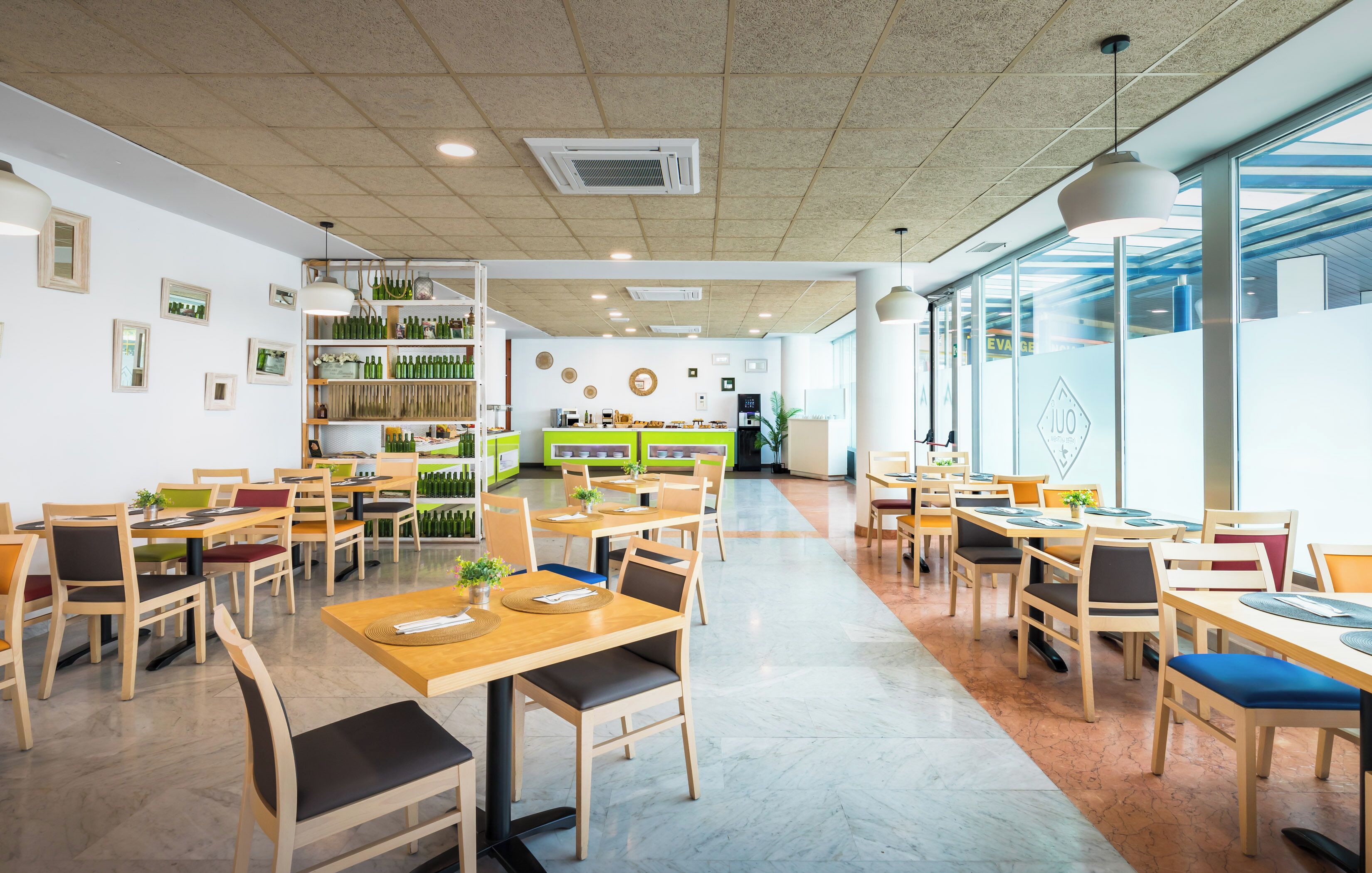 A spacious restaurant with wooden tables and chairs, a central counter, and a view through large windows.
