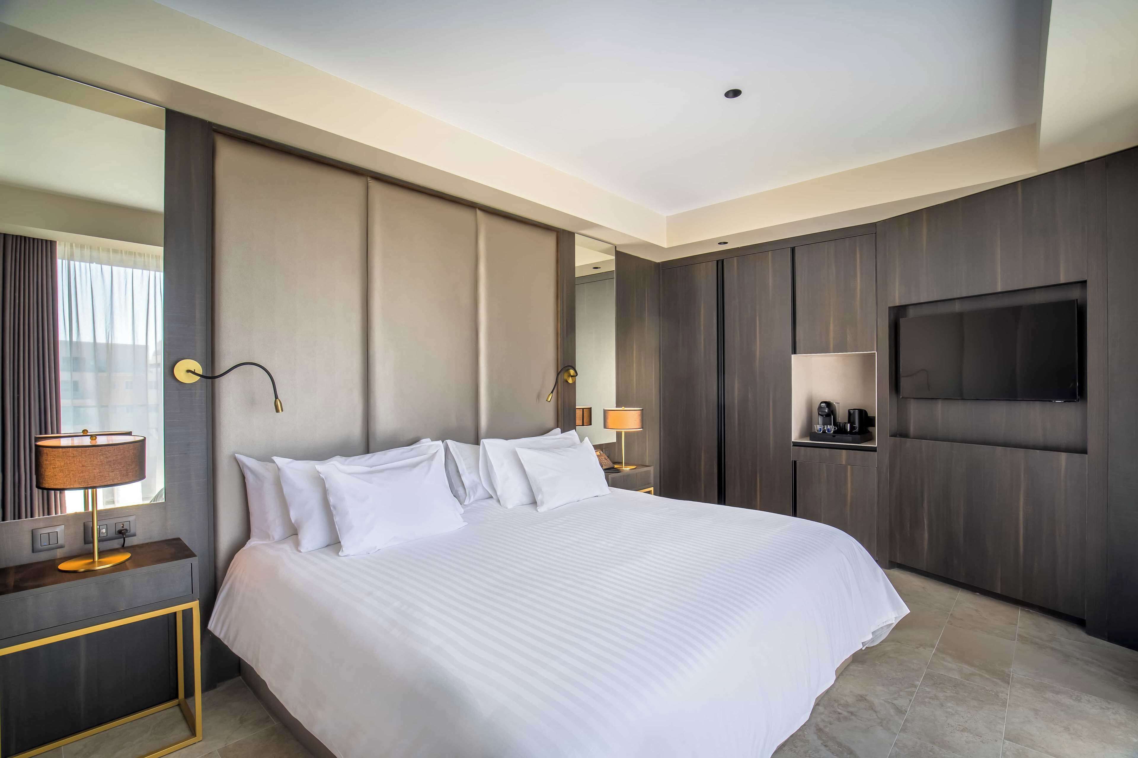 A modern hotel room with a large bed, a TV mounted on the wall, and a nightstand with a lamp.