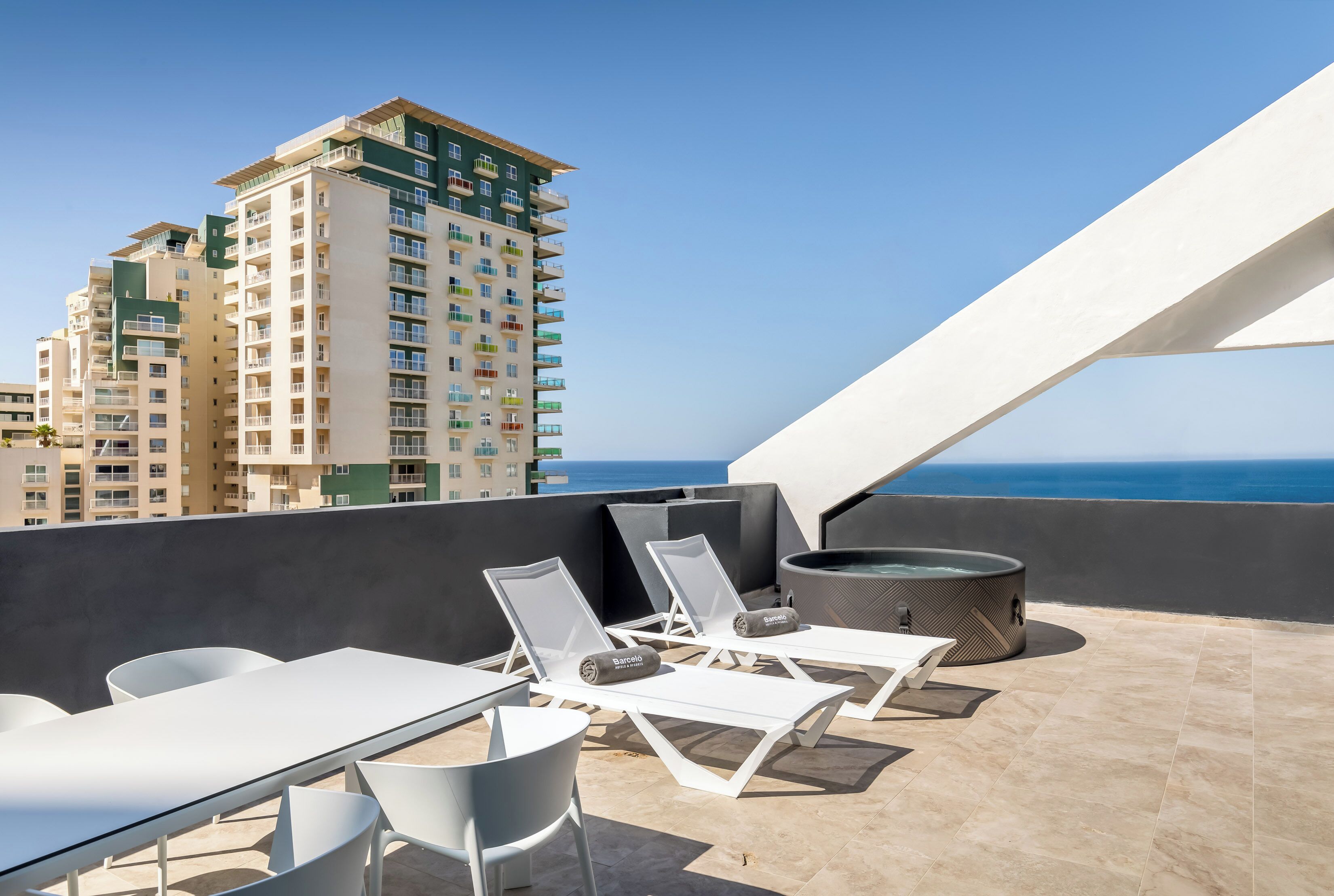 A rooftop terrace with white lounge chairs, a table, and a hot tub.