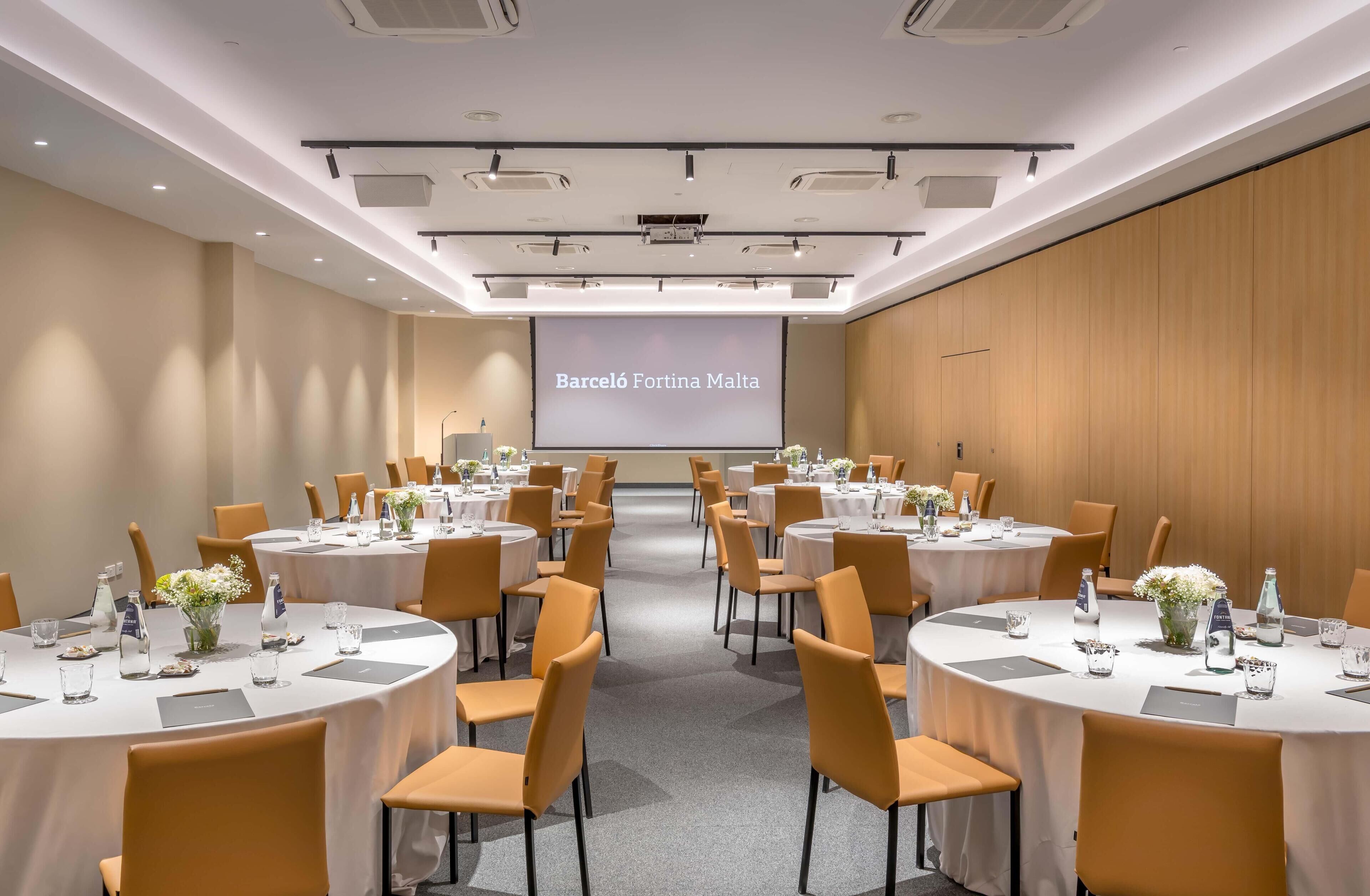 A conference room with multiple round tables, each with chairs, water bottles, and flower arrangements. A large screen displays 'Barceló Fortuna Malta'.