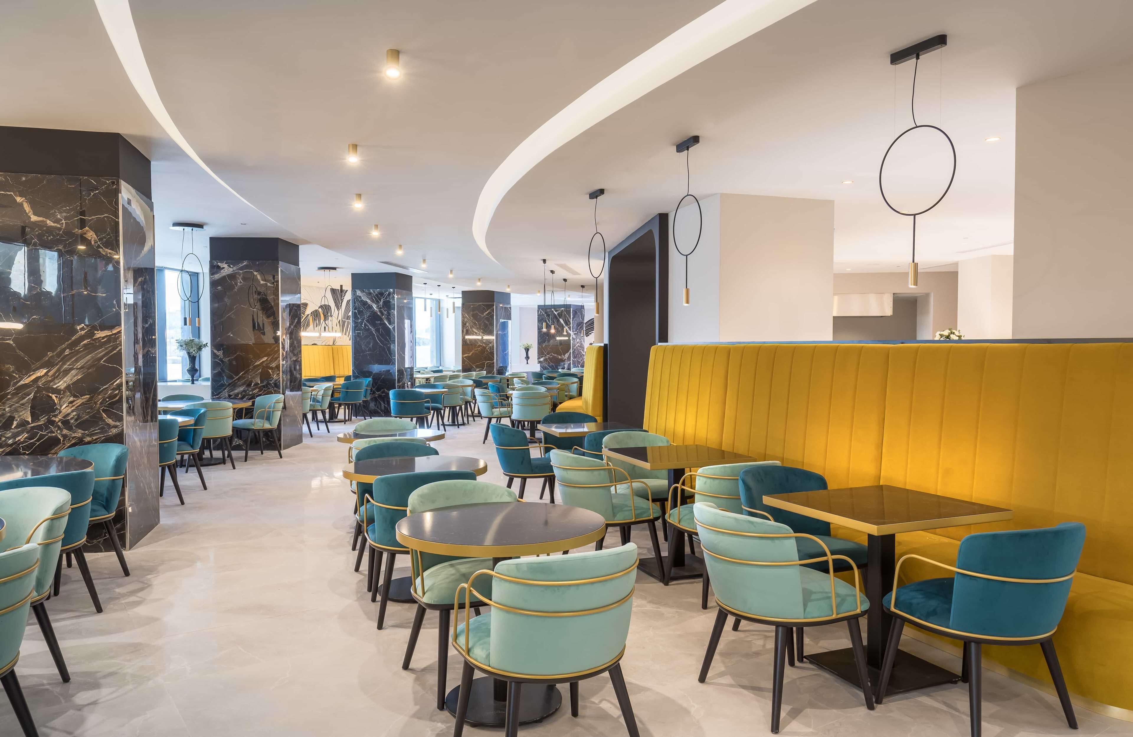 Un restaurante moderno con mesas redondas y sillas color turquesa, paredes de mármol y una pared de acento amarilla.