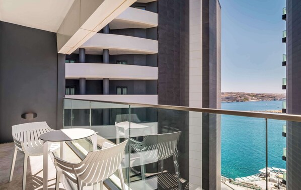 A balcony with white plastic chairs and tables, overlooking a body of water and a distant cityscape.