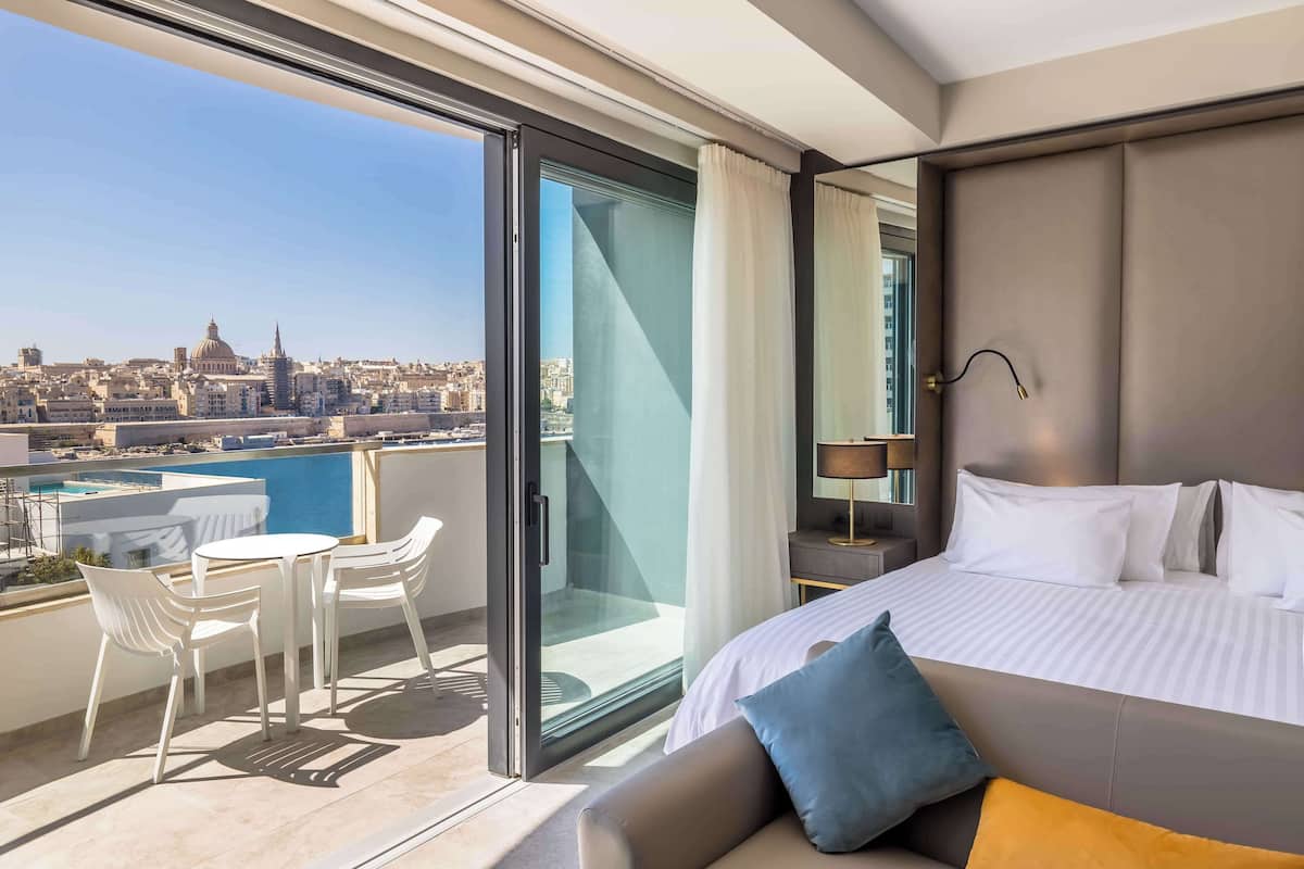 A hotel room with a bed, a nightstand, a lamp, a sofa, and a balcony with a table and chairs.
