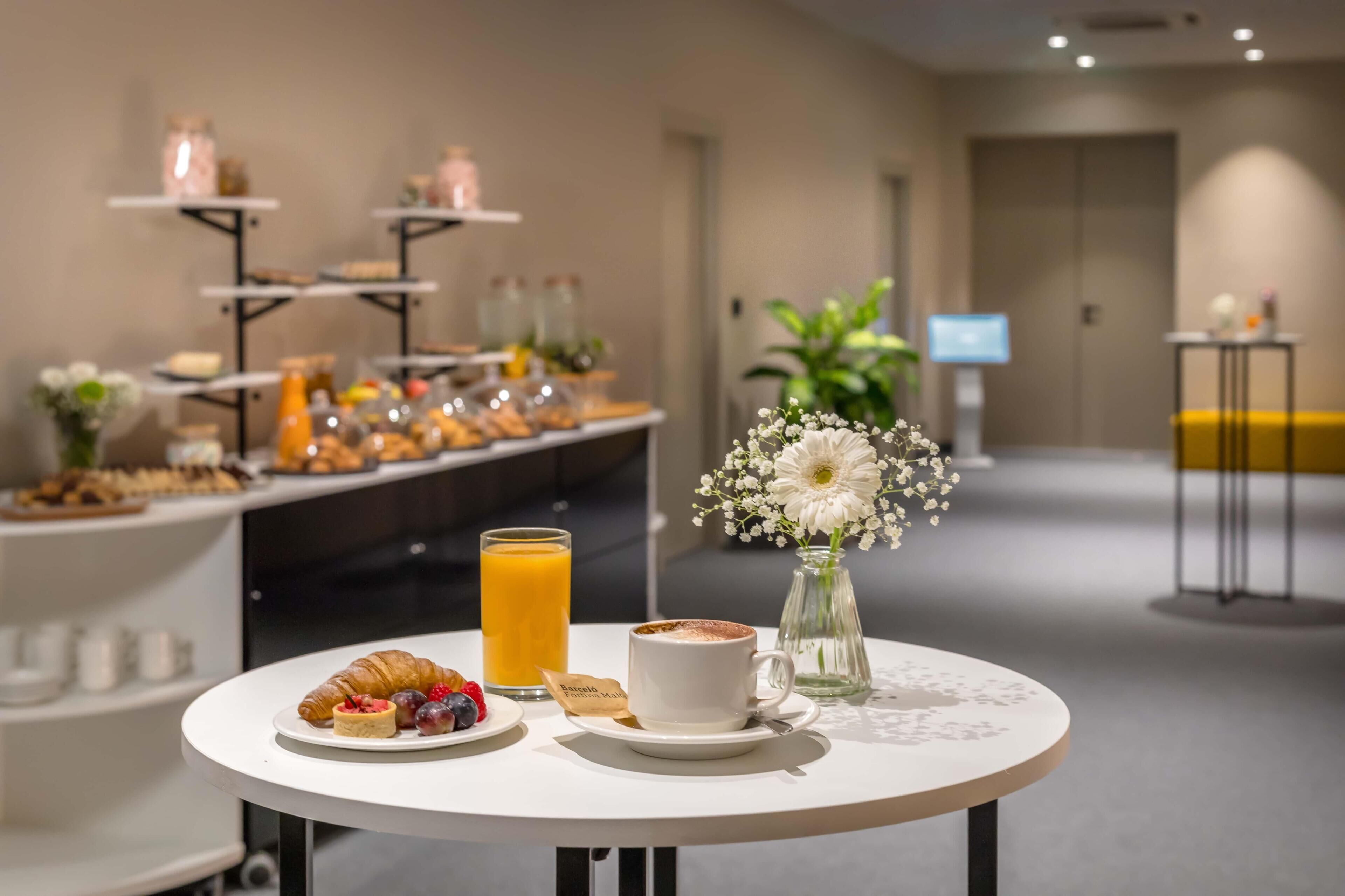 A breakfast setup with croissants, a cup of coffee, and a glass of orange juice on a table, with a buffet in the background.
