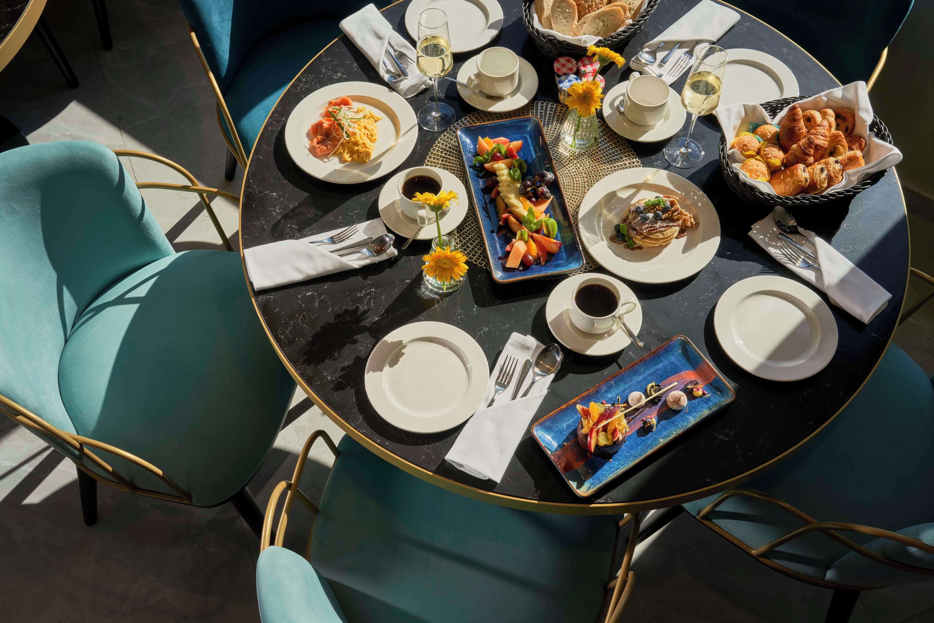 A round table with a variety of dishes, including croissants and fruit, and a small vase with sunflowers.