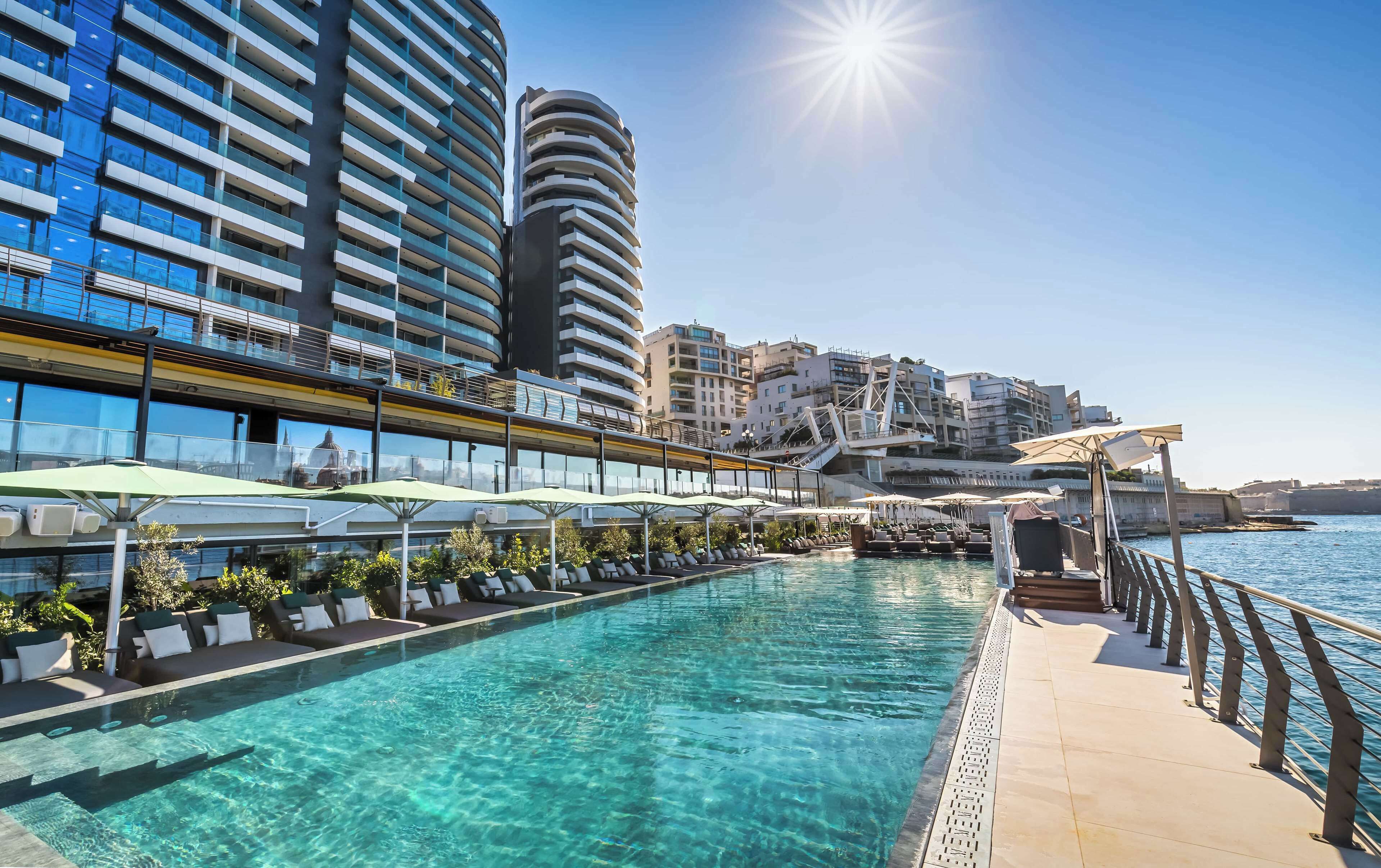 A poolside area with lounge chairs and umbrellas, a clear pool, and modern buildings in the background.