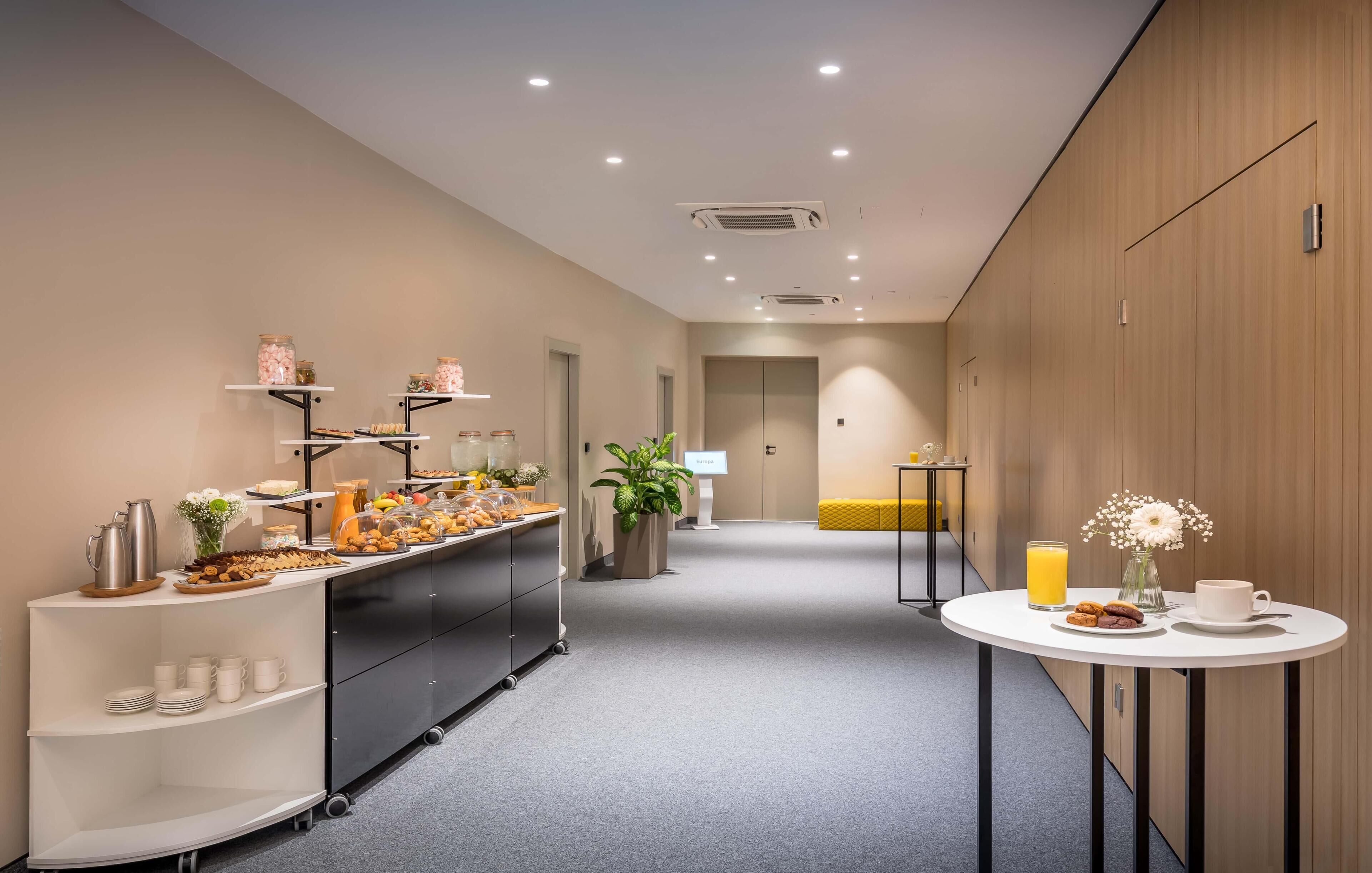 Un corredor moderno de hotel con una barra de desayuno, una zona de estar y una pequeña mesa con bebidas y pasteles.