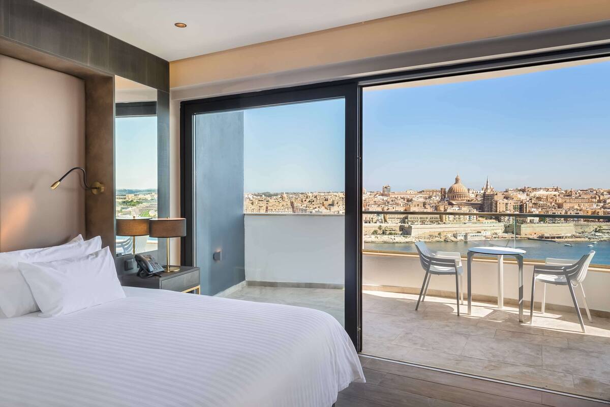 A hotel room with a large bed, a nightstand, a lamp, and a view of a cityscape through a balcony with a table and chairs.