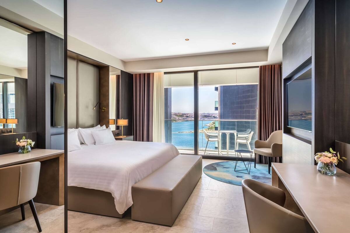 A modern hotel room with a large bed, a desk, a chair, a small table, and a balcony with a view of the sea.